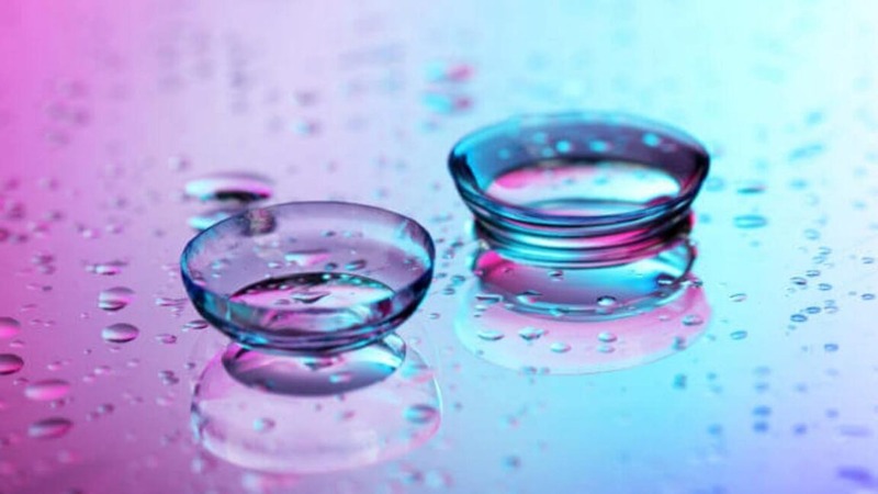 Two transparent contact lenses on a surface with water droplets, set against a pink and blue gradient background.